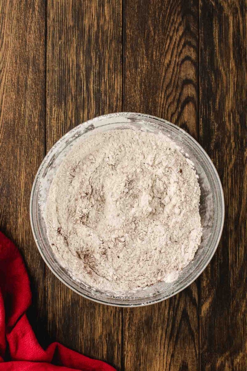 A glass bowl filled with a light brown dry flour mixture sits on a wooden surface next to a red cloth.