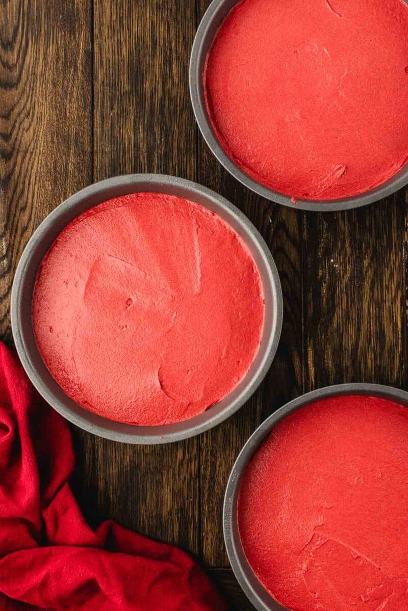 Three round baking pans filled with smooth, unbaked red cake batter sit on a wooden surface next to a red cloth.