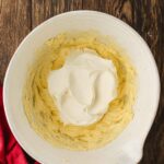 A white mixing bowl containing whipped yellow batter with a dollop of white cream on top, placed on a wooden surface next to a red cloth.