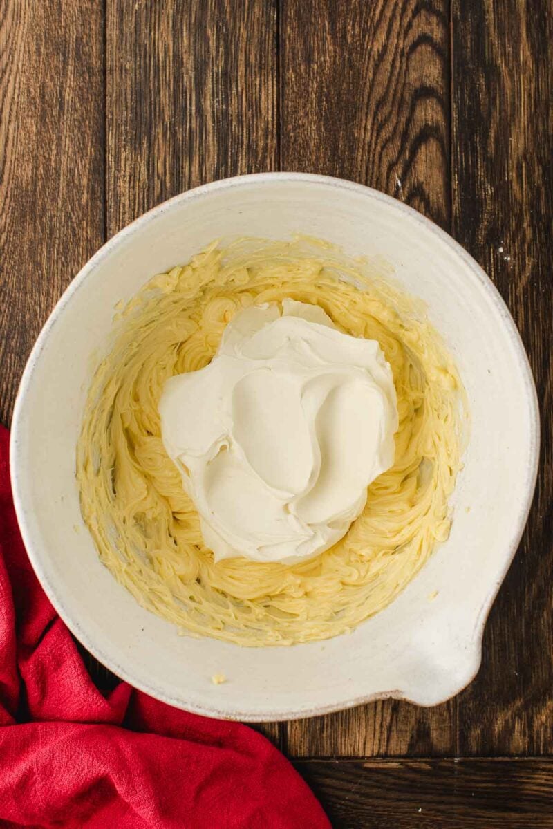 A white mixing bowl containing whipped yellow batter with a dollop of white cream on top, placed on a wooden surface next to a red cloth.