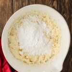 A white mixing bowl with yellow batter and a pile of powdered sugar on top, sitting on a wooden surface with a red cloth nearby.