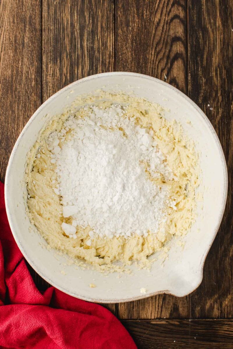 A white mixing bowl with yellow batter and a pile of powdered sugar on top, sitting on a wooden surface with a red cloth nearby.