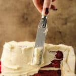 A hand uses an offset spatula to spread white frosting on a partially frosted red velvet cake on a plate.