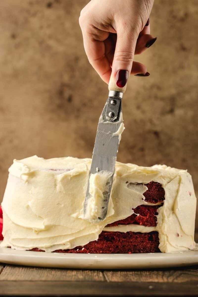 A hand uses an offset spatula to spread white frosting on a partially frosted red velvet cake on a plate.
