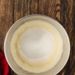 A metal mixing bowl with creamed butter and sugar sits on a wooden surface next to a red cloth.