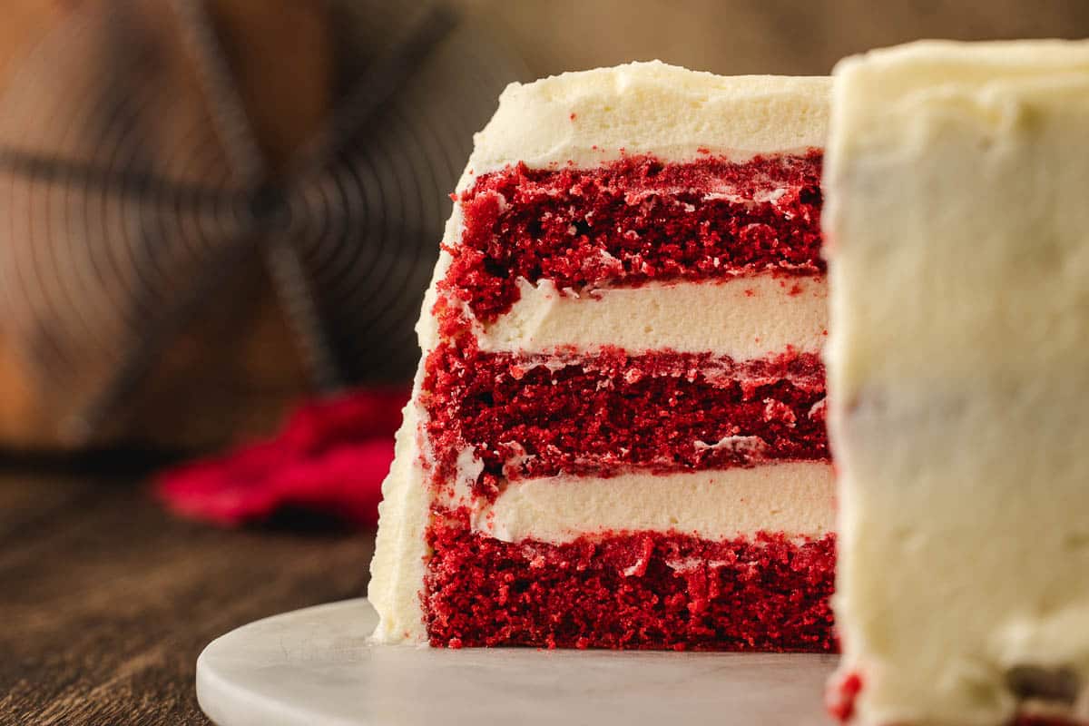 A close-up of a slice of red velvet cake with three layers of cream cheese frosting, sitting on a white plate.