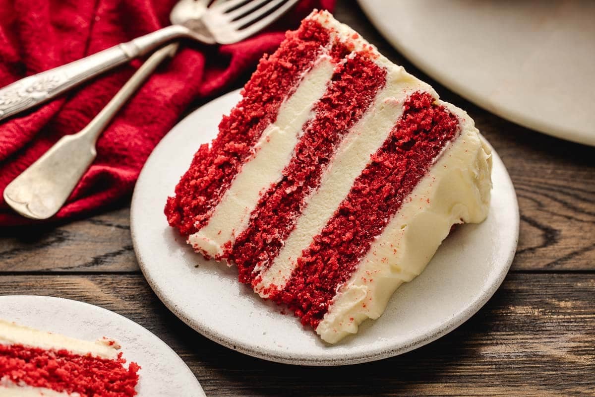 A slice of red velvet cake with cream cheese frosting on a white plate, with a fork and knife beside a red napkin on a wooden table.