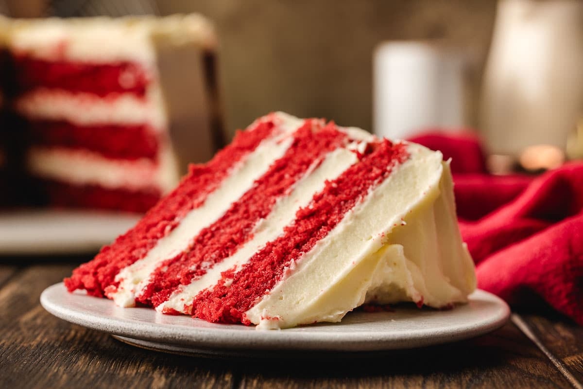 A slice of red velvet cake with four layers and cream cheese frosting sits on a plate, with the rest of the cake visible in the background.