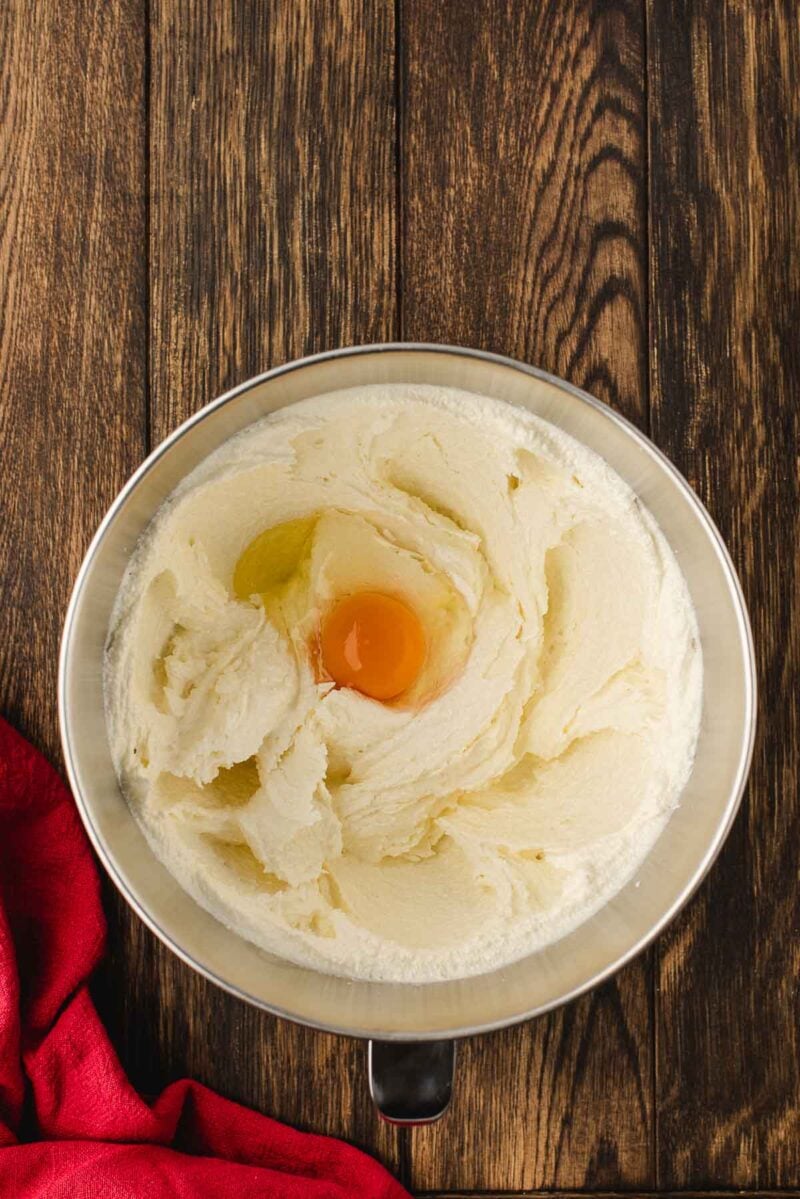 A metal mixing bowl with creamed butter and sugar, with an egg cracked in the center, sits on a wooden surface next to a red cloth.