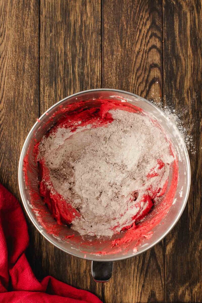 A metal mixing bowl with red batter and dry flour mixture on top sits on a wooden surface, next to a red cloth.