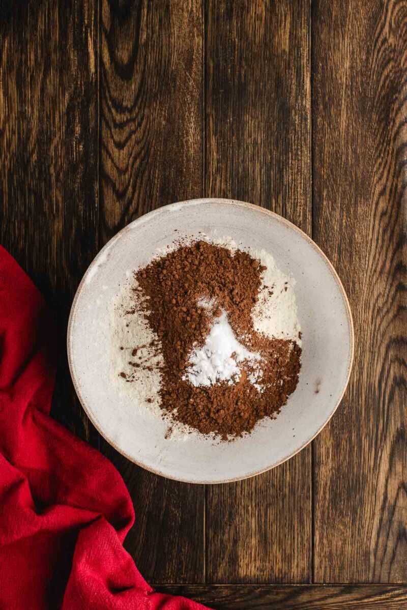 A white bowl containing flour, cocoa powder, and baking powder on a wooden surface with a red cloth to the side.