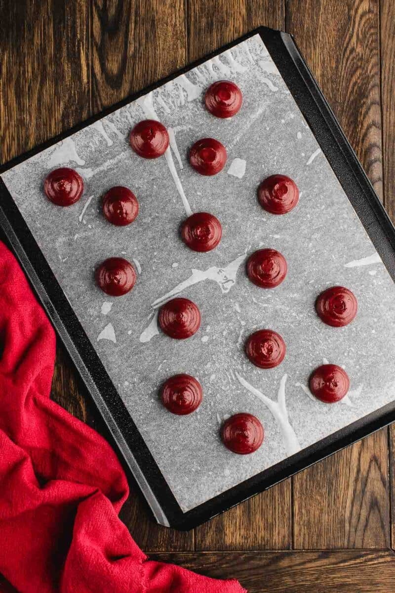 A baking tray lined with parchment paper holds fifteen evenly spaced, round blobs of red batter. A red cloth is placed beside the tray on a wooden surface.