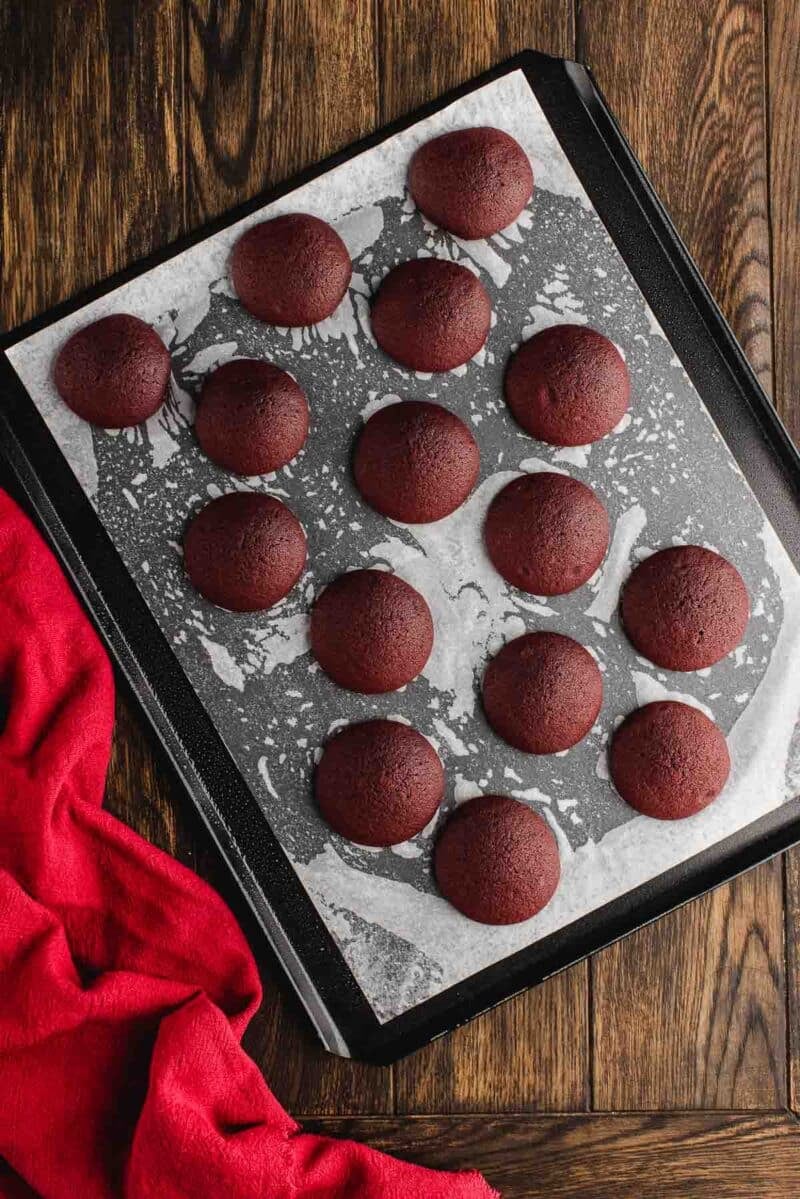 A baking tray lined with parchment paper holds twelve round, evenly spaced red velvet cookies. A red cloth is placed next to the tray on a wooden surface.