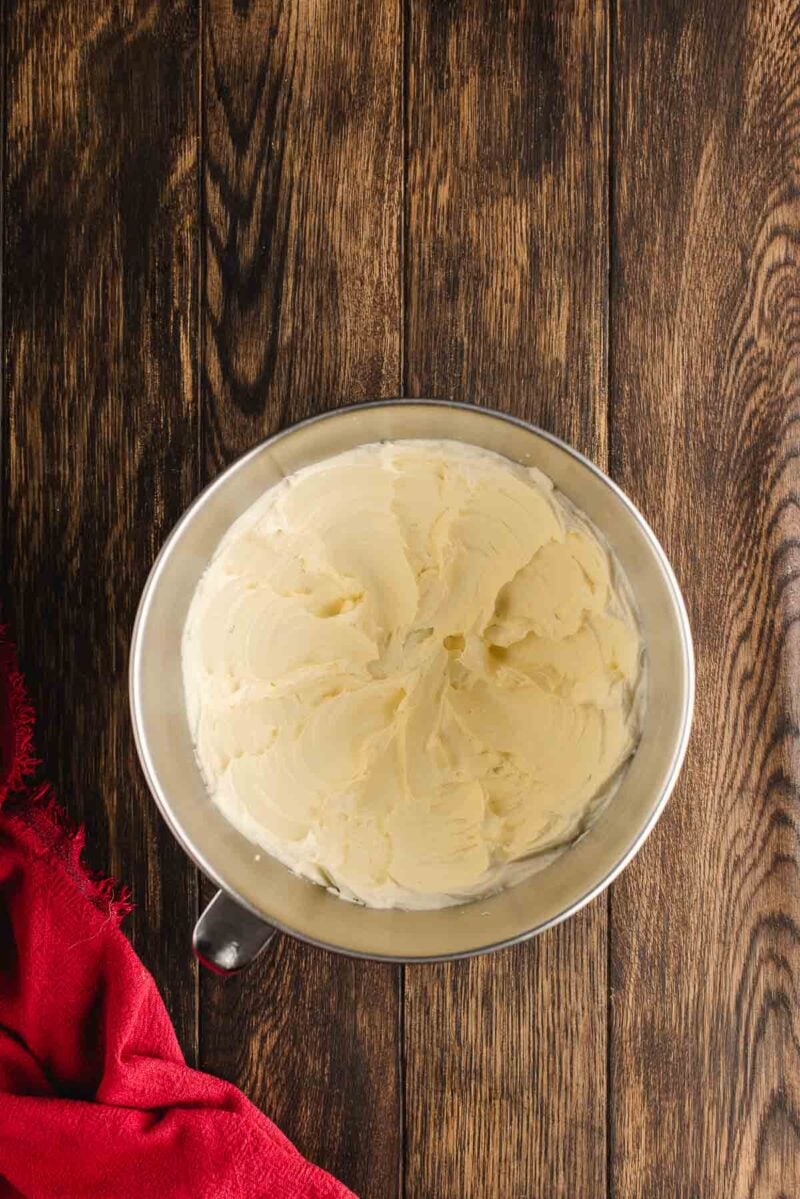 A metal mixing bowl filled with creamy, whipped butter sits on a wooden surface next to a red cloth.