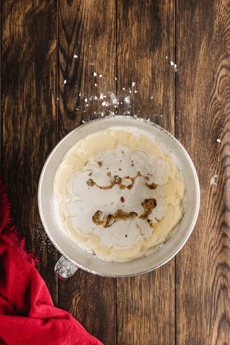 A metal mixing bowl with flour, brown sugar, and butter being mixed on a wooden surface, with a red cloth to the side.