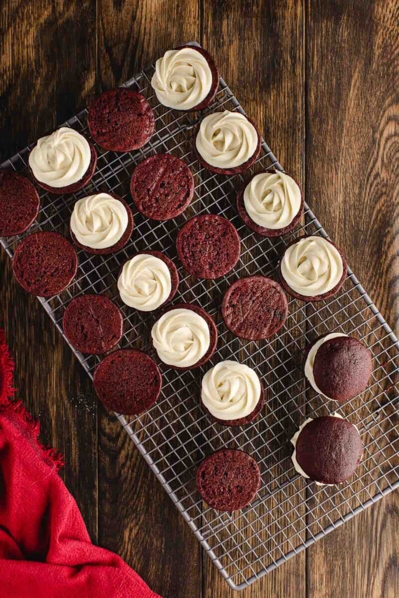A cooling rack holds several red velvet whoopie pies, some with visible white frosting filling, on a dark wooden surface with a red cloth in the corner.