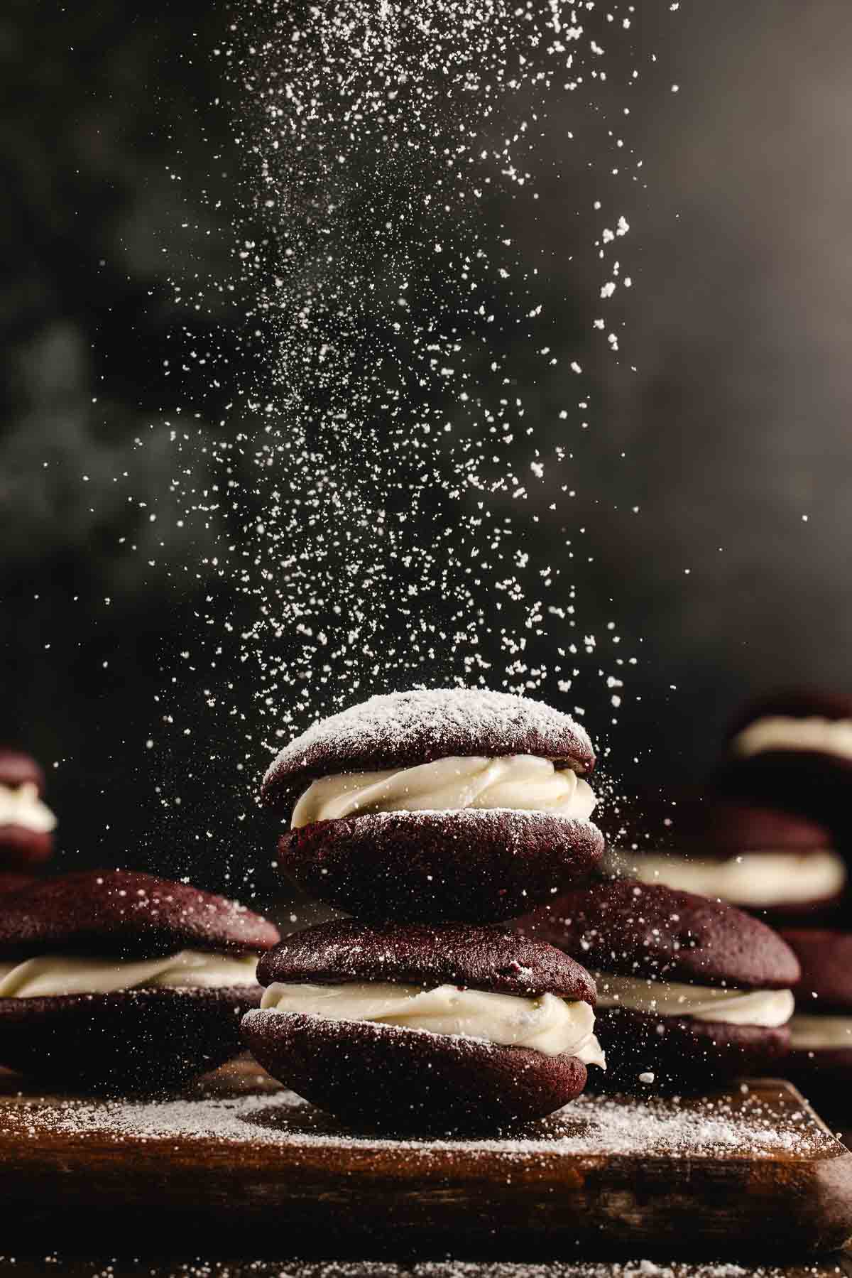 Three chocolate whoopie pies with cream filling stacked on a wooden surface, being dusted with powdered sugar, with more whoopie pies in the background.