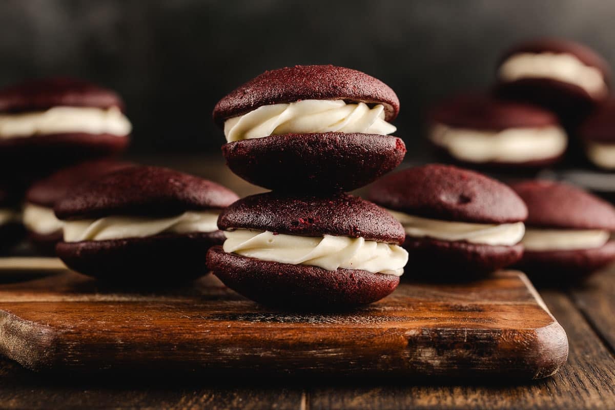 Two red velvet whoopie pies with white cream filling stacked on a wooden board, with more whoopie pies in the background.