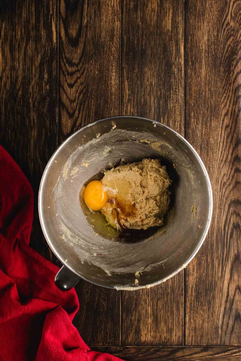 A metal mixing bowl with cookie dough, a raw egg, and vanilla extract sits on a wooden surface next to a red cloth.