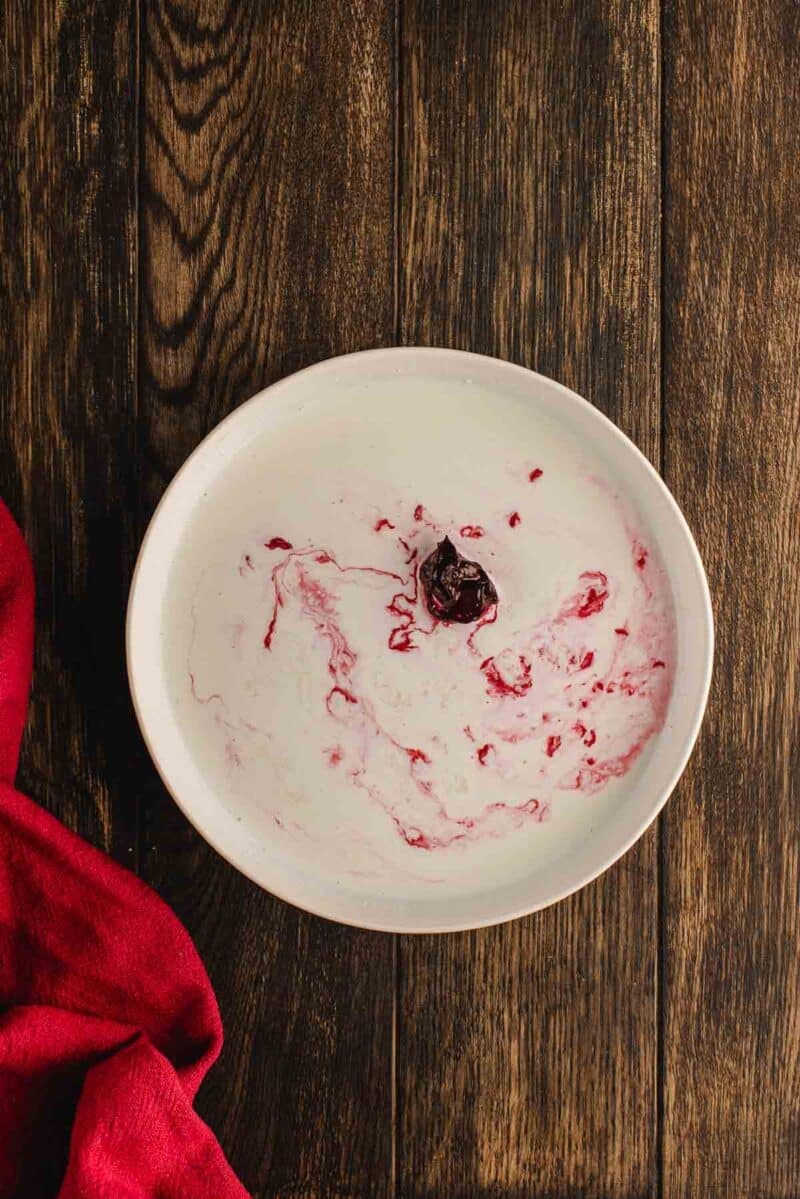 A white bowl with yogurt and a cherry on top, showing swirls of red, placed on a wooden surface next to a red cloth.