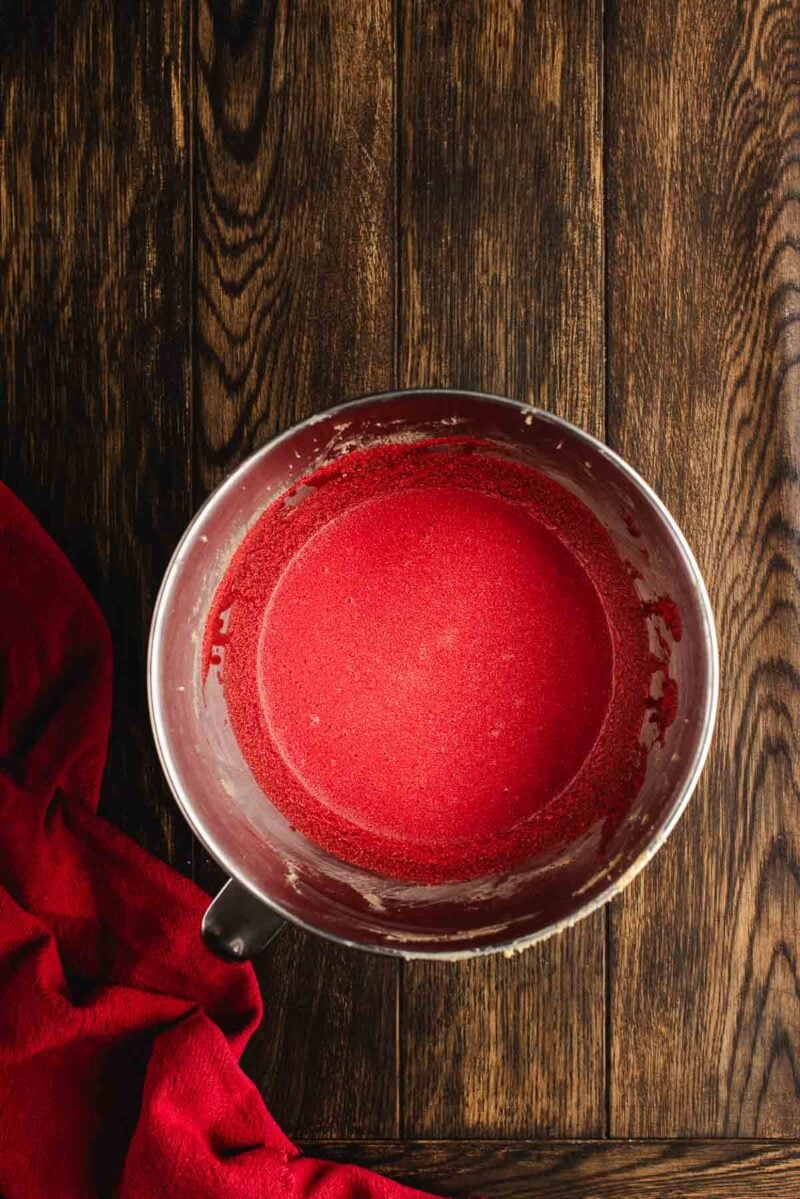 A metal mixing bowl filled with smooth red batter sits on a dark wooden surface next to a red cloth.