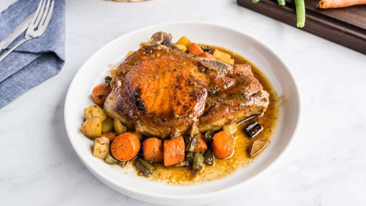 A cooked pork chop sits on a white plate, served with gravy and assorted roasted vegetables including carrots, green beans, and potatoes.