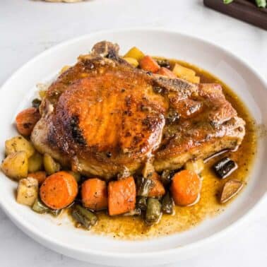 A cooked pork chop sits on a white plate, served with gravy and assorted roasted vegetables including carrots, green beans, and potatoes.