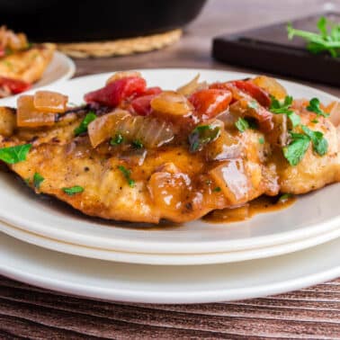 A cooked chicken breast topped with a tomato, onion, and herb sauce is served on a white plate, with a wooden board and fresh herbs in the background.