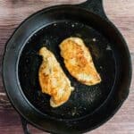 Two cooked chicken breasts in a black cast iron skillet on a wooden surface.