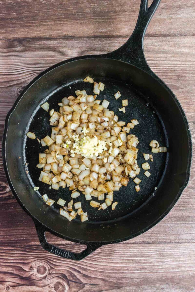 Diced onions and minced garlic are sautéing in a black cast iron skillet on a wooden surface.