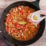 A hand pours liquid from a measuring cup into a cast iron skillet filled with diced vegetables, tomatoes, and a bay leaf on a wooden surface.