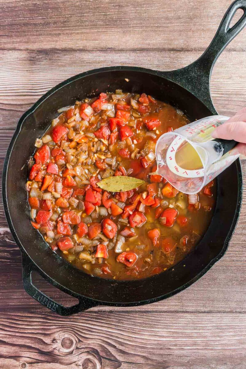 A hand pours liquid from a measuring cup into a cast iron skillet filled with diced vegetables, tomatoes, and a bay leaf on a wooden surface.