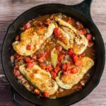 Three cooked chicken breasts with tomatoes, onions, and a bay leaf in a cast iron skillet on a wooden surface.