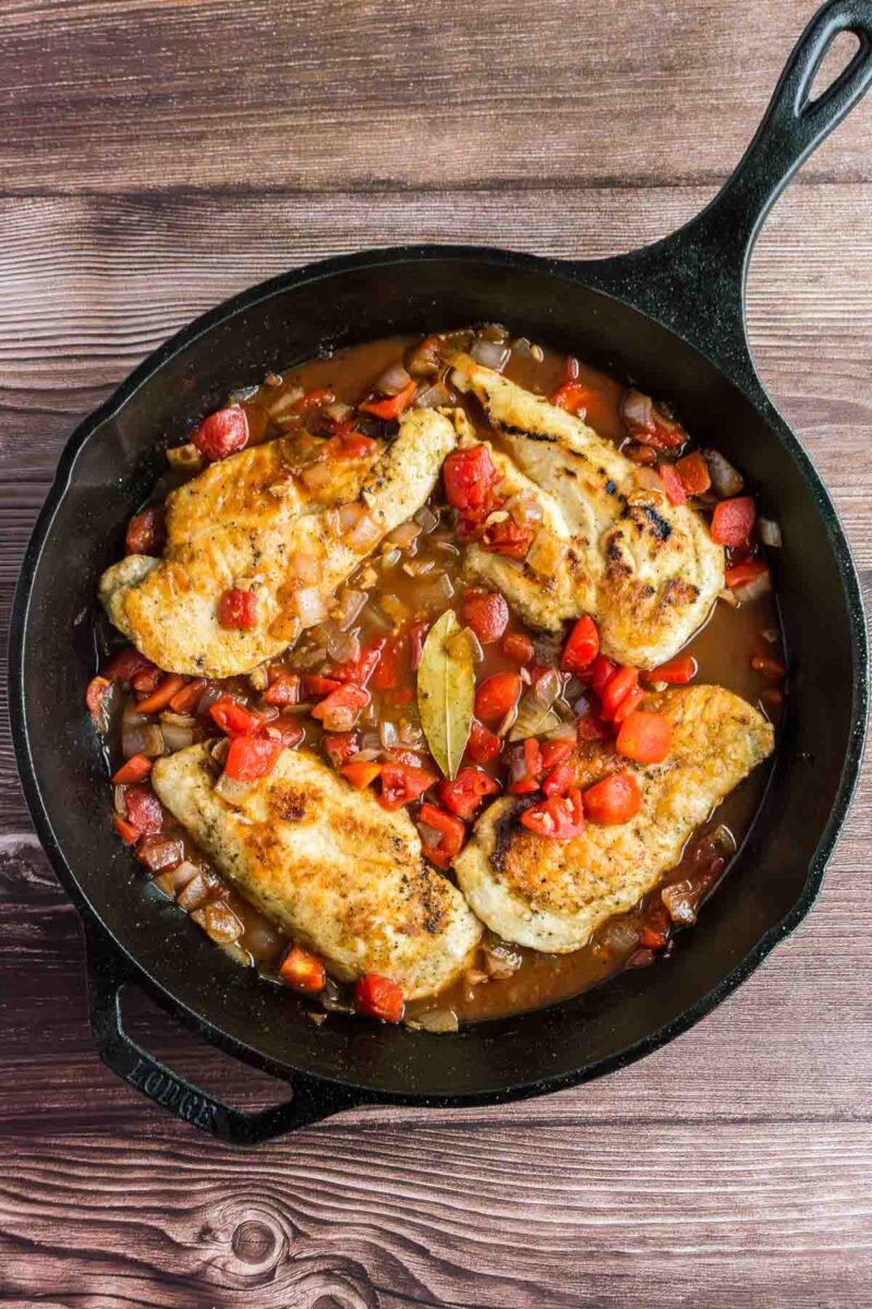 Three cooked chicken breasts with tomatoes, onions, and a bay leaf in a cast iron skillet on a wooden surface.