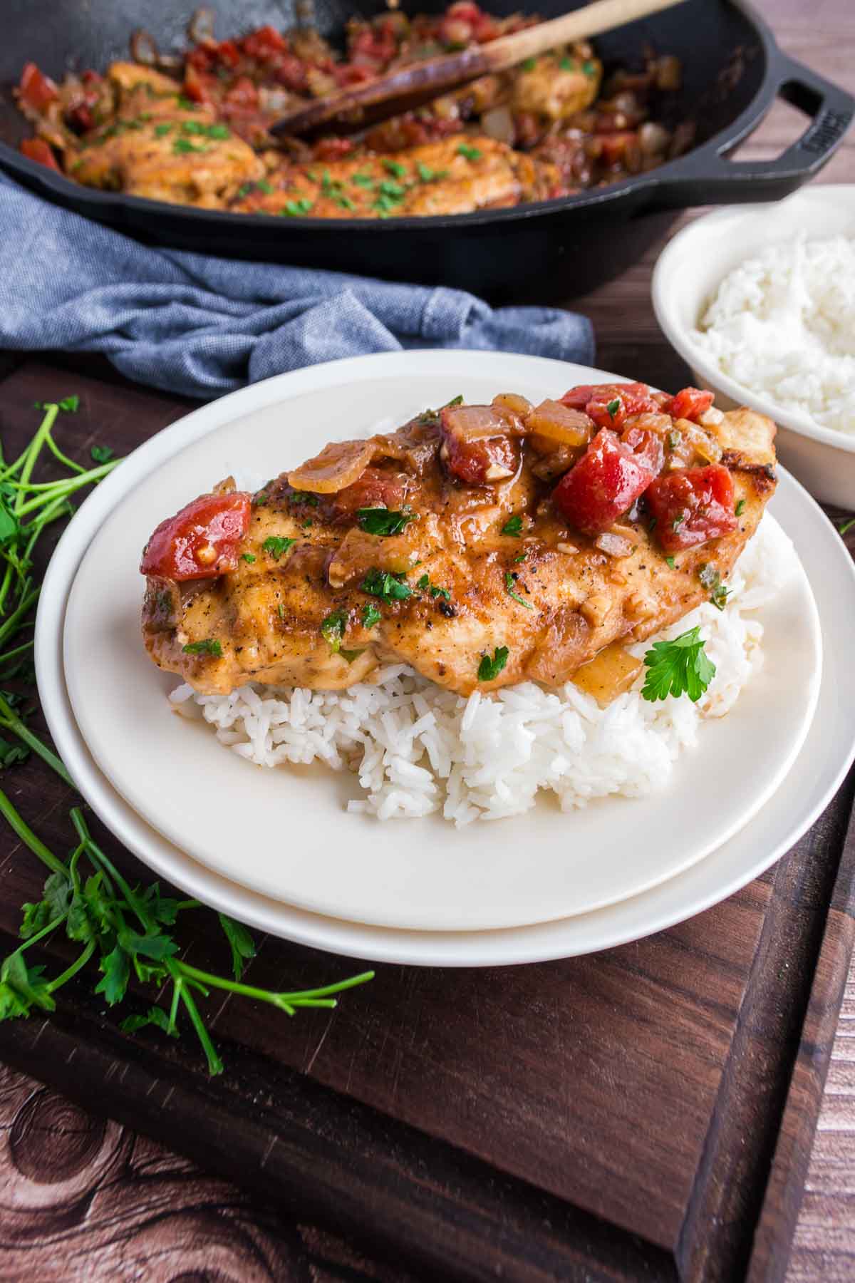 Grilled chicken breast with tomato and onion sauce served over white rice on a plate, with skillet and a bowl of rice in the background.