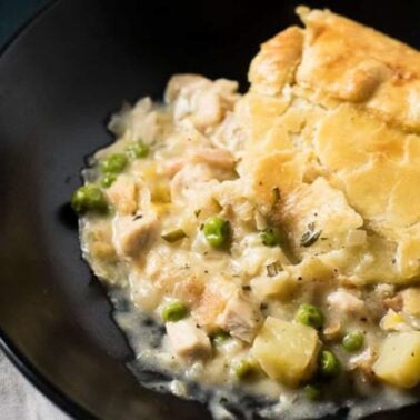 A serving of chicken pot pie with golden crust, filled with savory protein, peas, potatoes, and rich gravy in a creamy sauce, on a black plate.