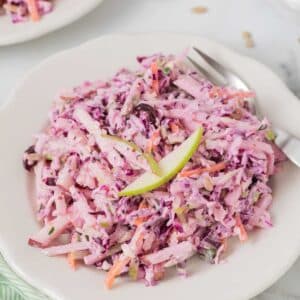 A plate of creamy apple coleslaw made with shredded cabbage and carrots, garnished with two thin slices of green apple, served with a fork.