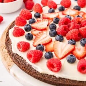 A dessert pizza with a brownie crust, white frosting, and topped with fresh raspberries, blueberries, and sliced strawberries on a wooden board.