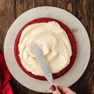 A hand uses an offset spatula to spread white frosting on a round red velvet cake layer on a marble board, set on a wooden surface.