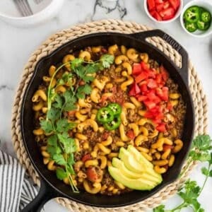 A cast iron skillet filled with macaroni, ground meat, tomatoes, and garnished with sliced avocado, chopped tomatoes, jalapeño, and cilantro, on a woven trivet.