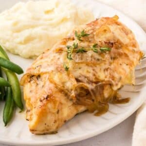 A plate with french onion chicken topped with herbs, served with mashed potatoes and green beans.
