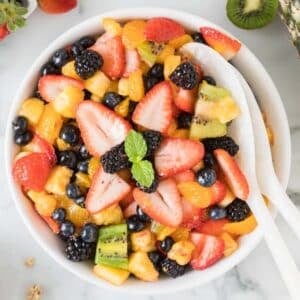 A bowl of mixed fruit salad with strawberries, blueberries, blackberries, kiwi, and orange segments, garnished with mint and served with white utensils.