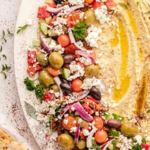 A plate with hummus drizzled with olive oil, served alongside a Greek salad with olives, cherry tomatoes, cucumber, red onion, feta, and herbs.