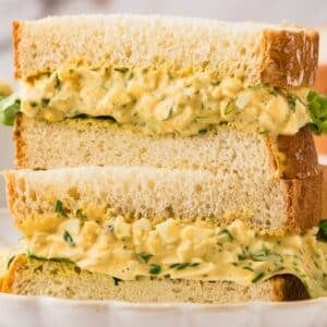 A close-up of an egg salad sandwich made with thick slices of white bread, stacked halves showing creamy filling with chopped herbs and lettuce.