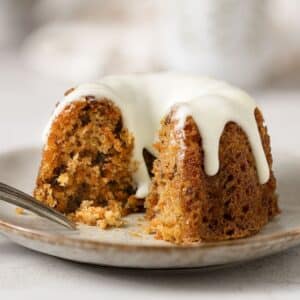 A mini carrot cake with cream cheese icing sits on a plate, partially sliced with a fork.