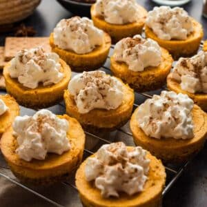 Mini pumpkin cheesecakes topped with whipped cream and a sprinkle of cinnamon are arranged on a cooling rack.