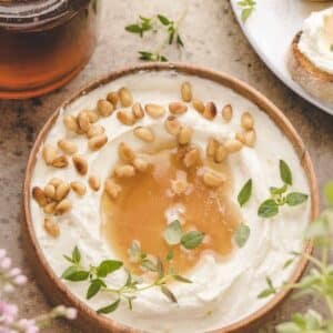 A wooden bowl of whipped ricotta topped with honey, pine nuts, and fresh herbs, placed on a stone surface.