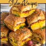 A platter of breakfast sliders with eggs, cheese, and bacon on poppy seed buns, garnished with herbs and served with grapes.