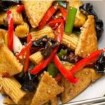 A bowl of stir-fried tofu with baby corn, red bell peppers, green onions, and black mushrooms, garnished with sesame seeds for extra flavorโone of the tastiest tofu recipes you'll savor.