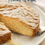 A sliced sweet almond cake topped with powdered sugar and crunchy toasted nuts is displayed on a white plate with a serving utensil.